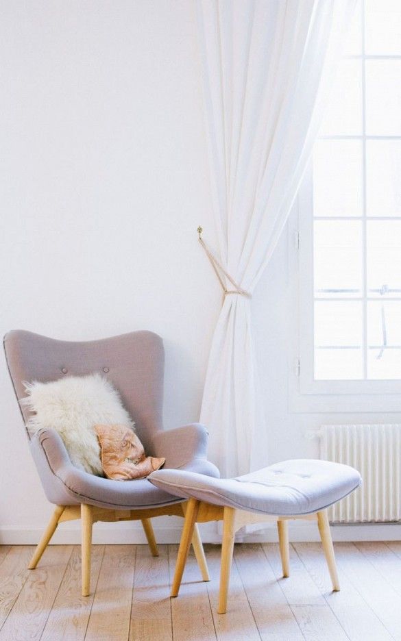 Matching grey fabric armchair and foot stool with fur pillow by a large  window with all white curtains.